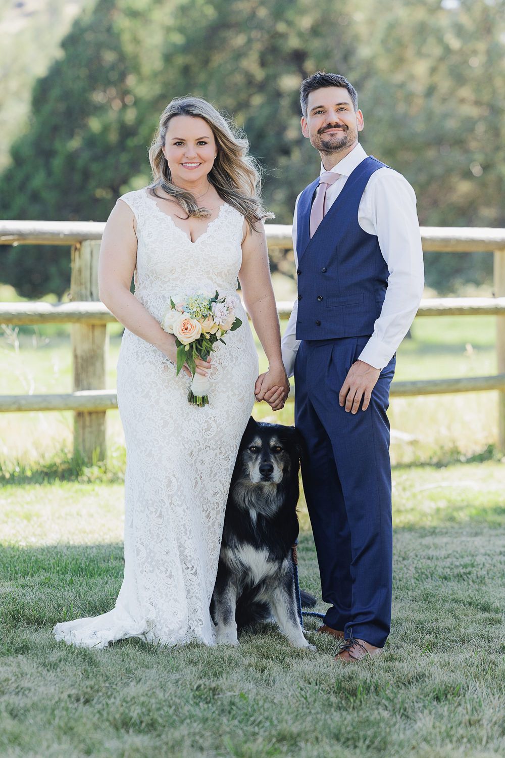 Newlywed couple poses with their black German Shepherd dog in front of a rustic wooden fence in a grassy field.