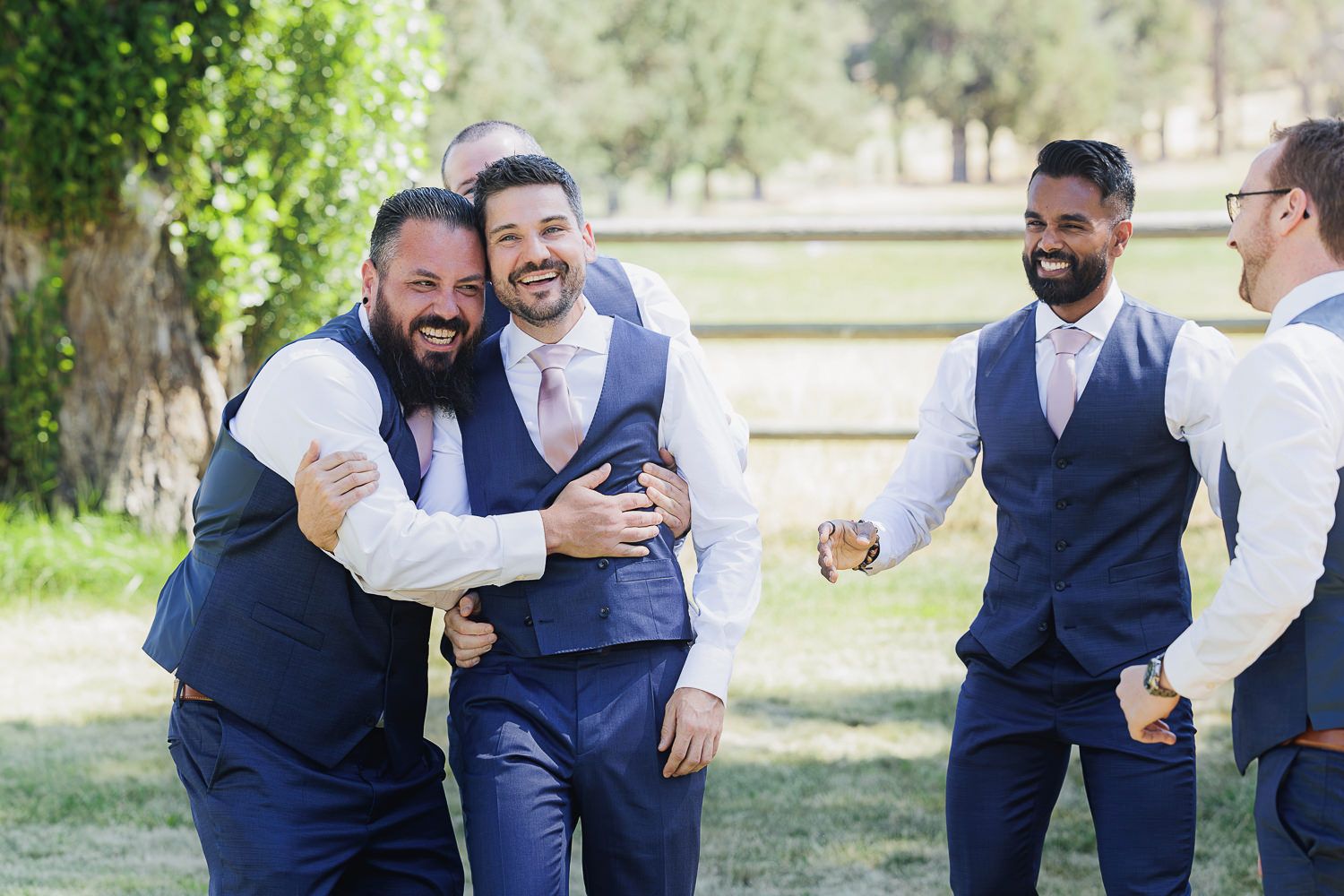 Groomsmen in navy blue vests and suits laughing and embracing outdoors during a wedding celebration.