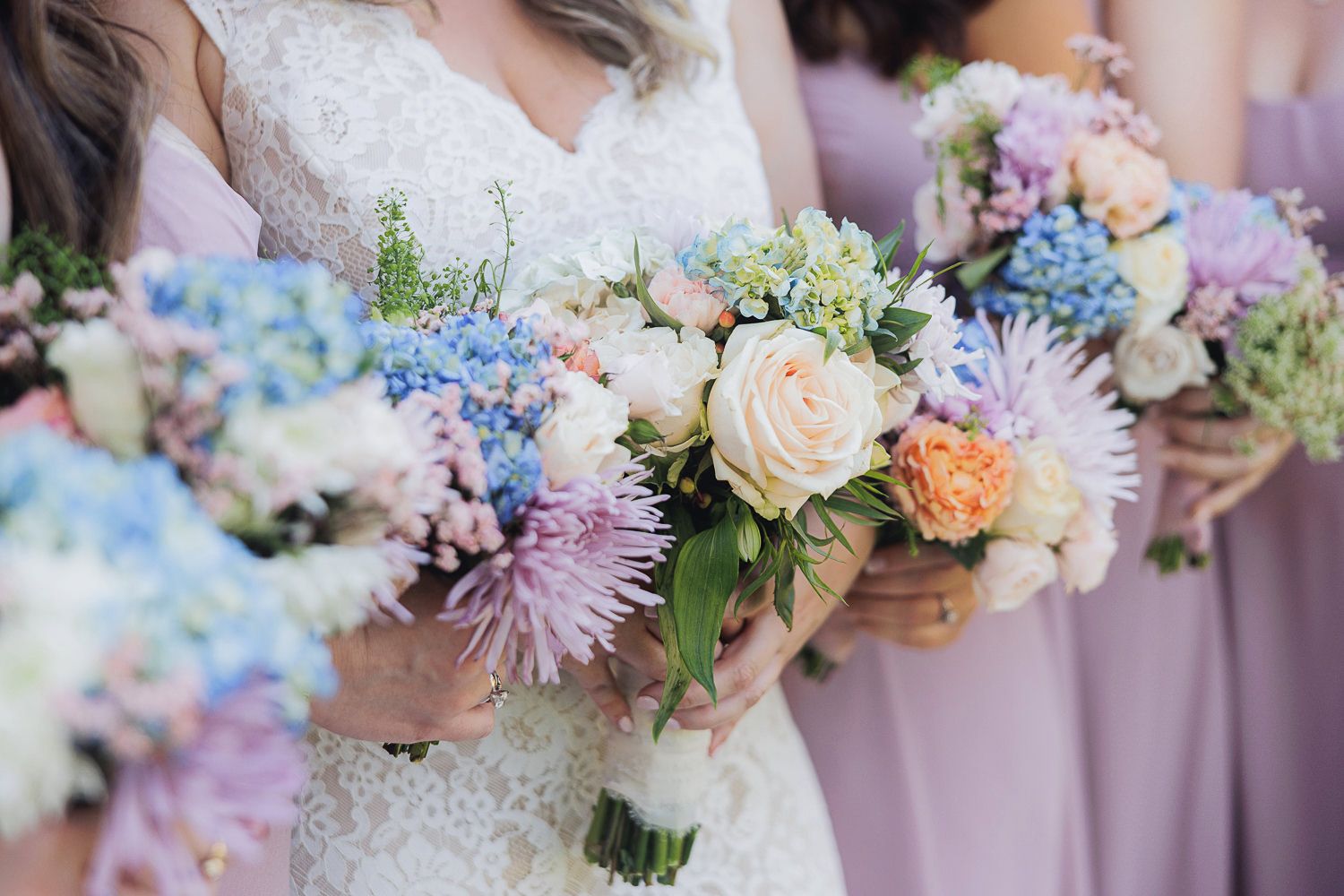 Close-up of wedding bouquets with white roses, blue hydrangeas, and pink dahlias against mauve bridesmaid dresses.