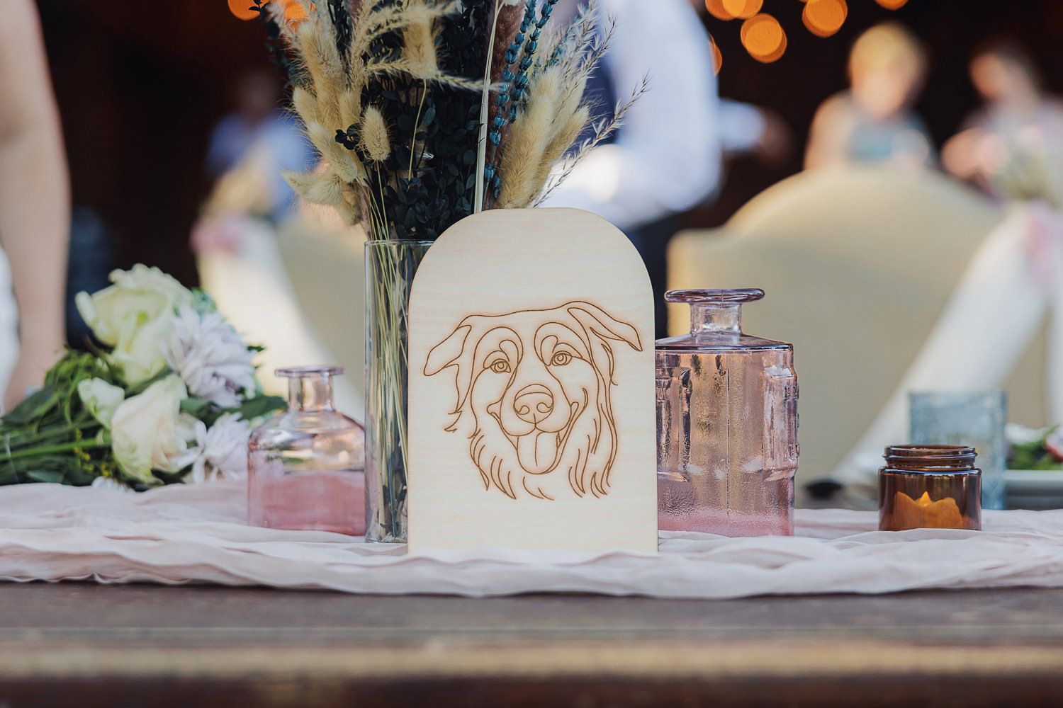 A rustic table setting with a dog-themed luminous candle holder, pink glassware, and white floral centerpiece.