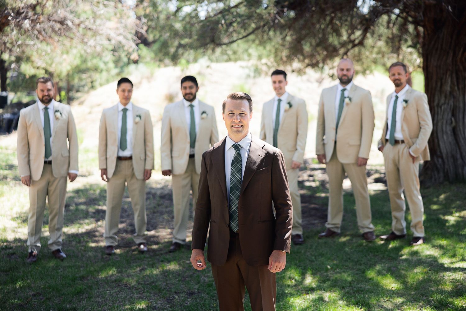 Groom in brown suit stands in front of groomsmen wearing light khaki suits and blue ties outdoors on sunny day.