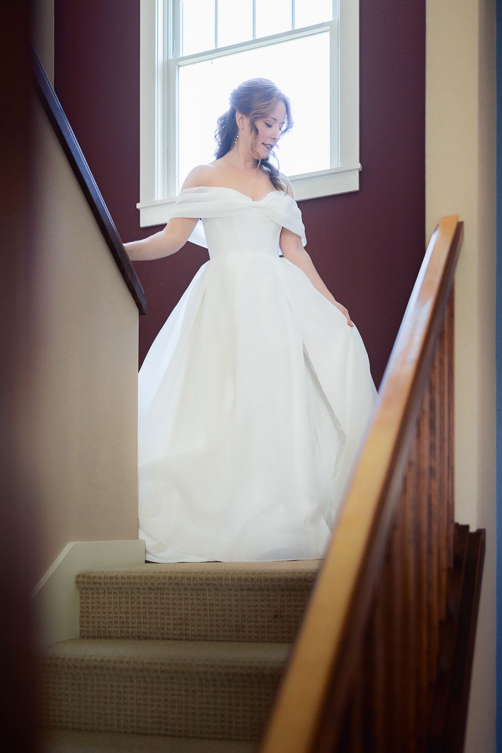 A bride in an elegant off-shoulder white ballgown stands gracefully on a carpeted staircase by a bright window.