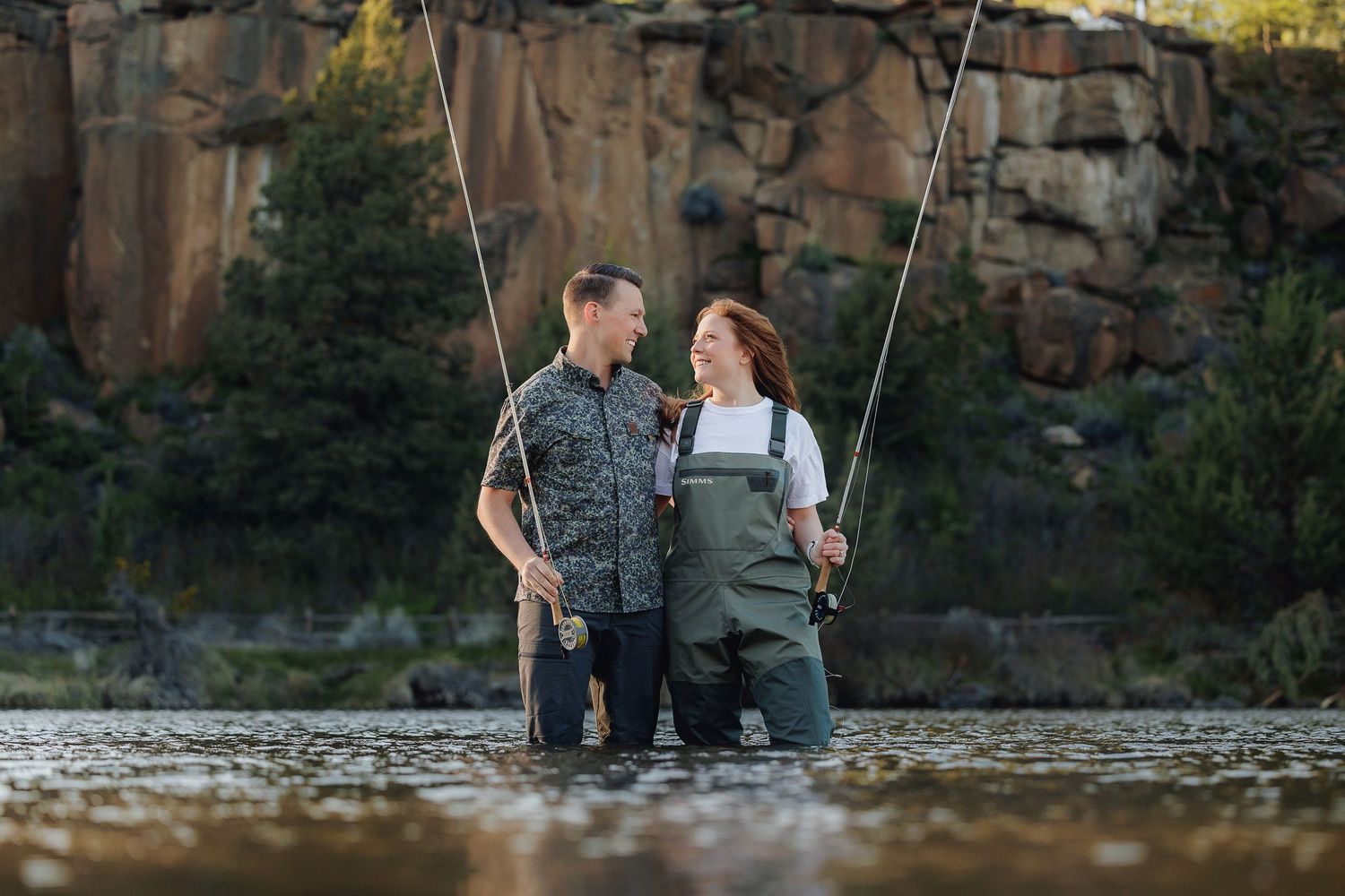 Two people in fishing waders stand together in a stream holding fishing rods with cliffs in the background.