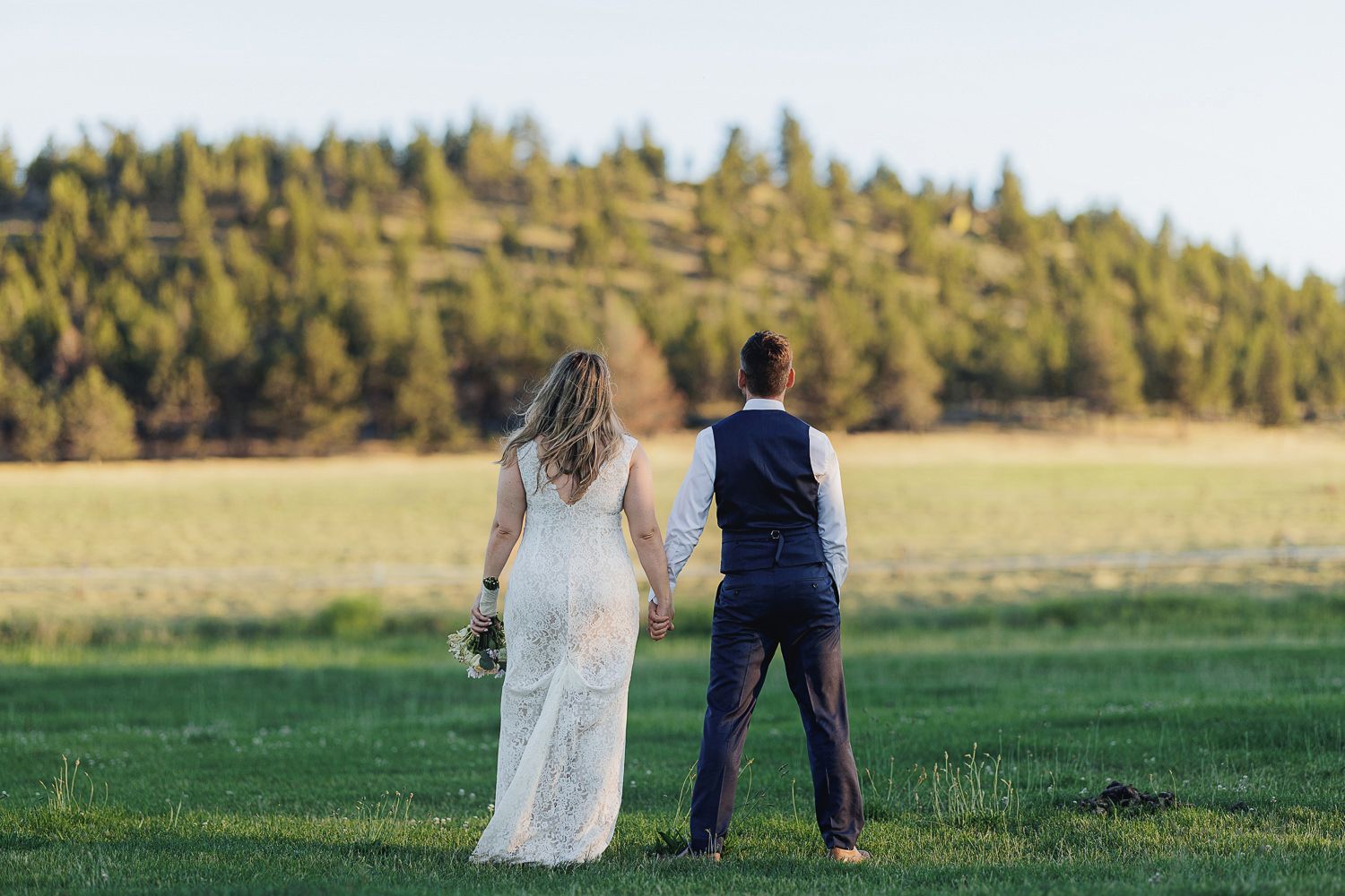 A couple in wedding attire stand together in a grassy field with golden hills in the background at sunset.