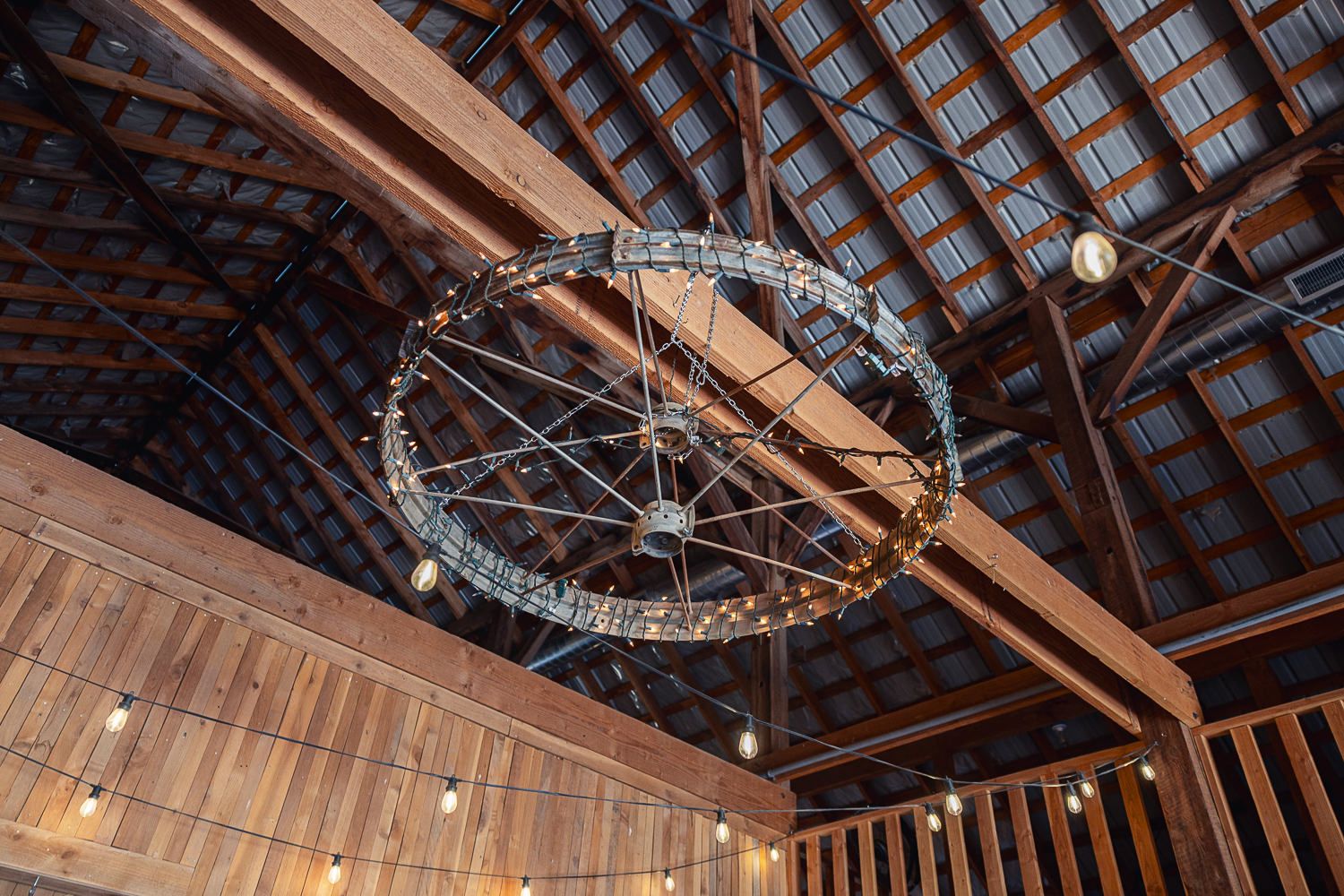 Rustic exposed wooden beam ceiling with industrial lighting fixtures and dangling string lights creates a barn-like atmosphere.
