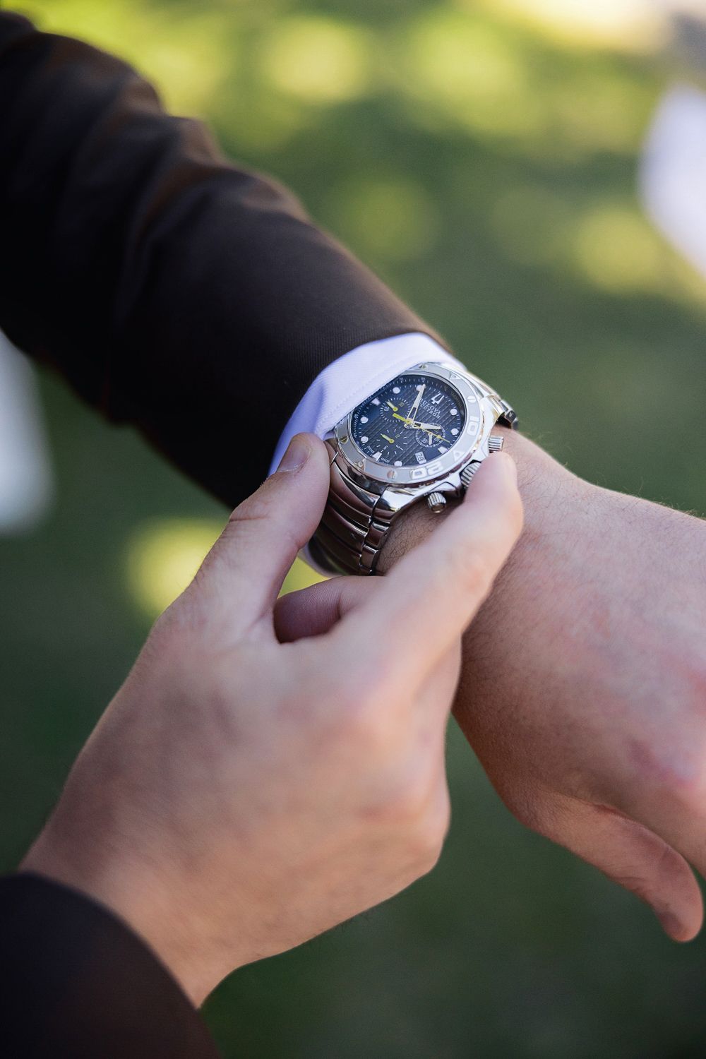 Two people wearing similar steel chronograph watches compare their timepieces in a natural outdoor setting.