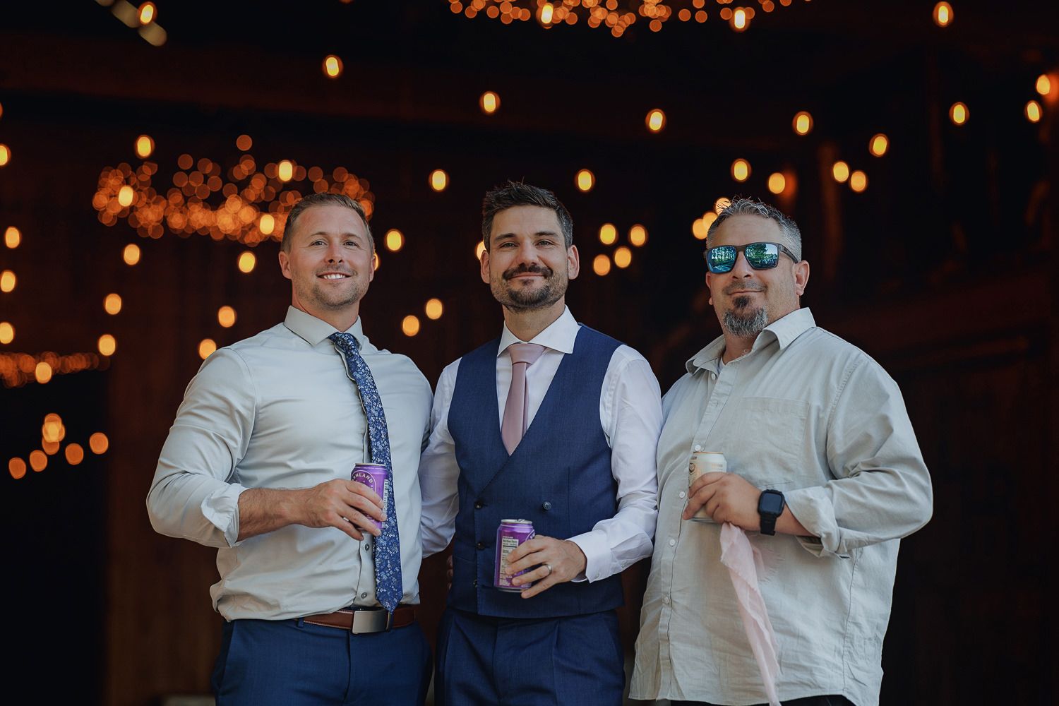 Groomsmen laughing and enjoying themselves at a wedding reception with twinkling lights in the background.
