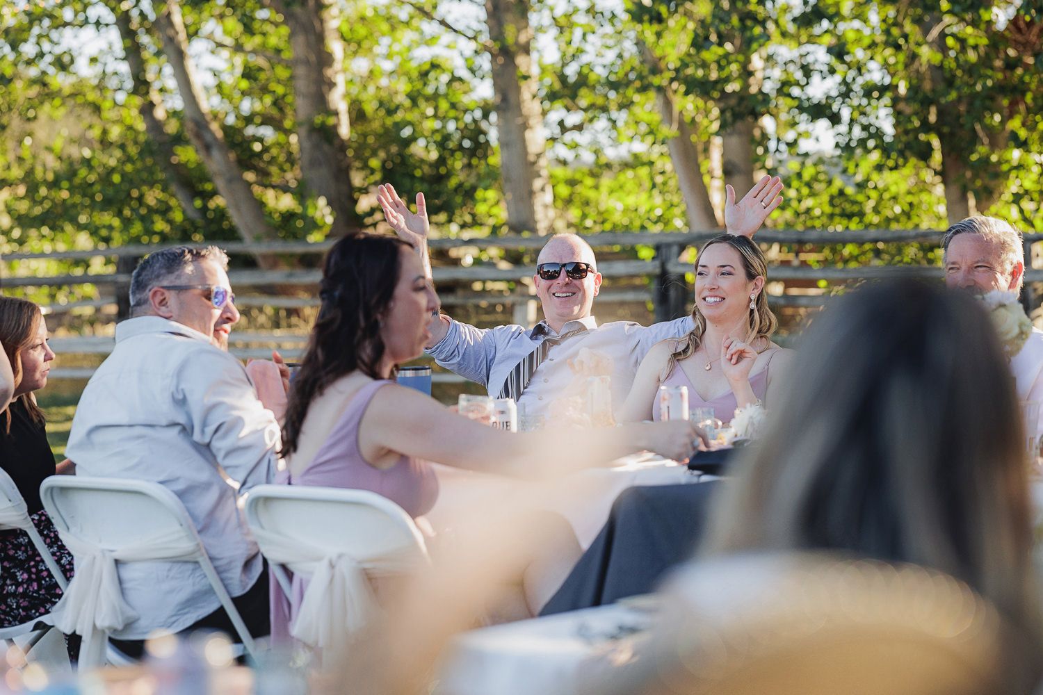 People gathered outdoors at white tables enjoying a casual dining event in sunny weather with birch trees in background.
