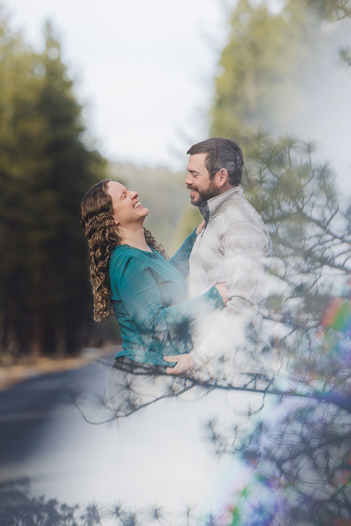 A romantic outdoor couple portrait through blurred tree branches with dreamy lighting and soft bokeh effects.