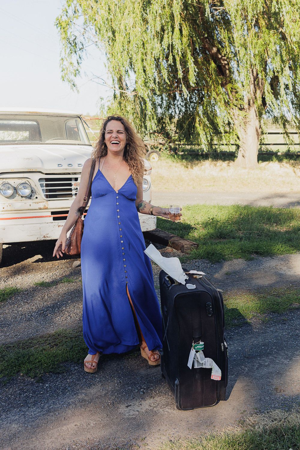 Someone in a flowing blue dress sits playfully near suitcases under a willow tree on a sunny day.