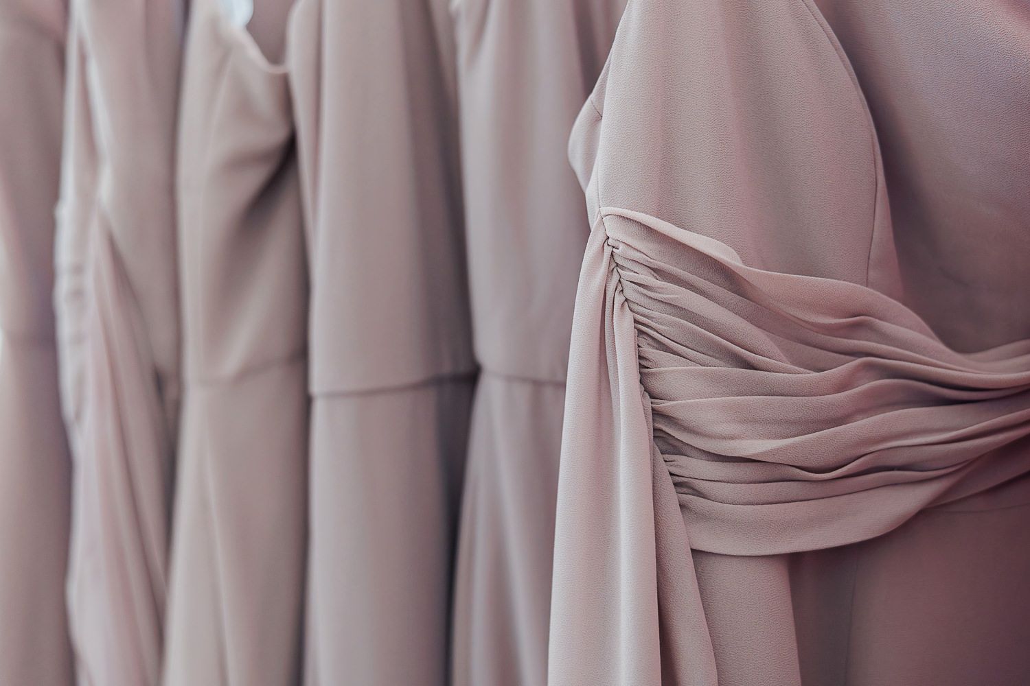 Several dusty rose colored bridesmaid dresses with ruched detailing hanging on a clothing rack.