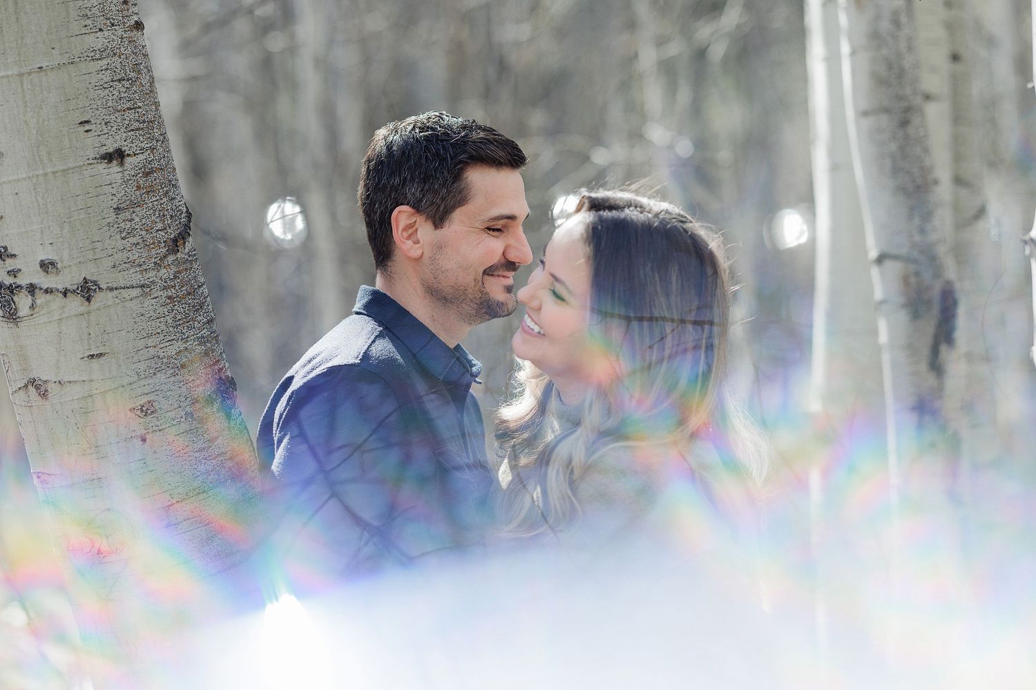 A romantic winter engagement photo with rainbow lens flares in a snowy aspen forest.