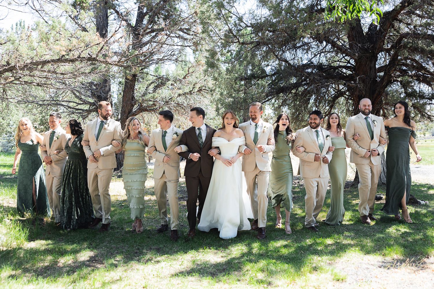Wedding party in neutral khaki and sage green attire poses together under trees in outdoor garden setting.