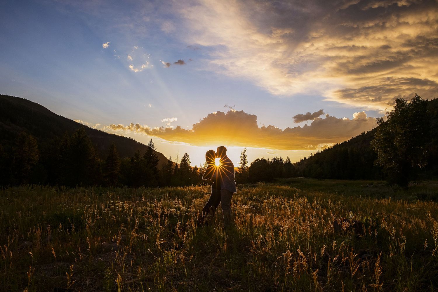 Silhouetted couple in grassy meadow with golden sunset and mountains creating dramatic mountain landscape.
