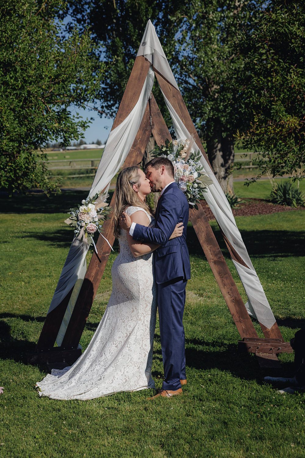 Wedding couple embraces under a decorated wooden triangle arch with draped fabric and floral arrangements.