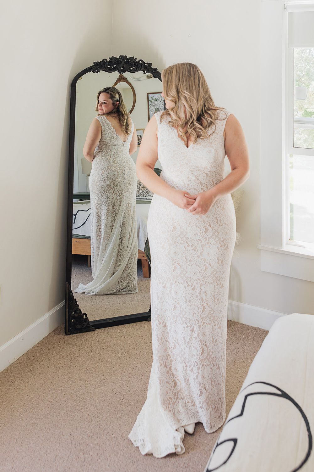 Elegant full-length white lace wedding gown reflection in ornate black vintage mirror in bright dressing room setting.