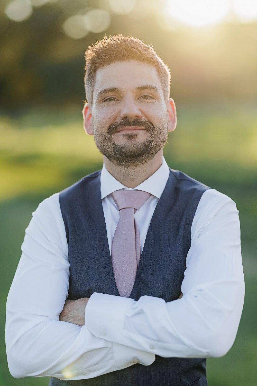 Wedding day portrait of a groom in a navy vest and pink tie standing outside at sunset.