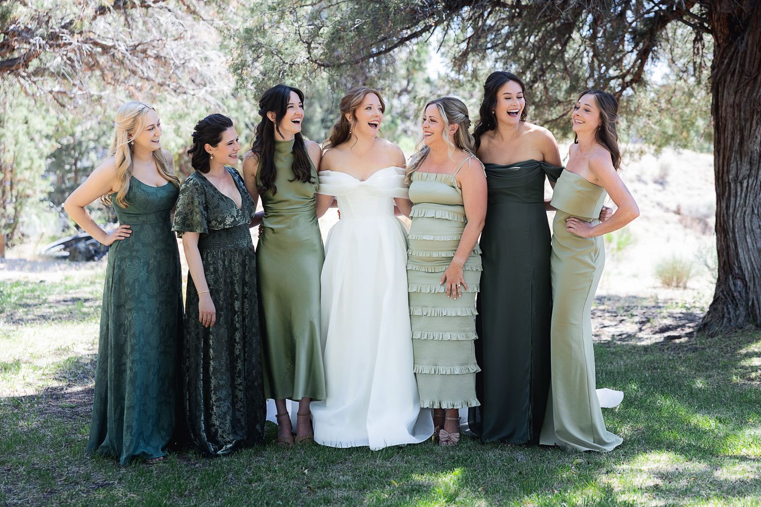 Wedding party in green and white dresses pose under trees in an outdoor setting during spring or summer celebration.