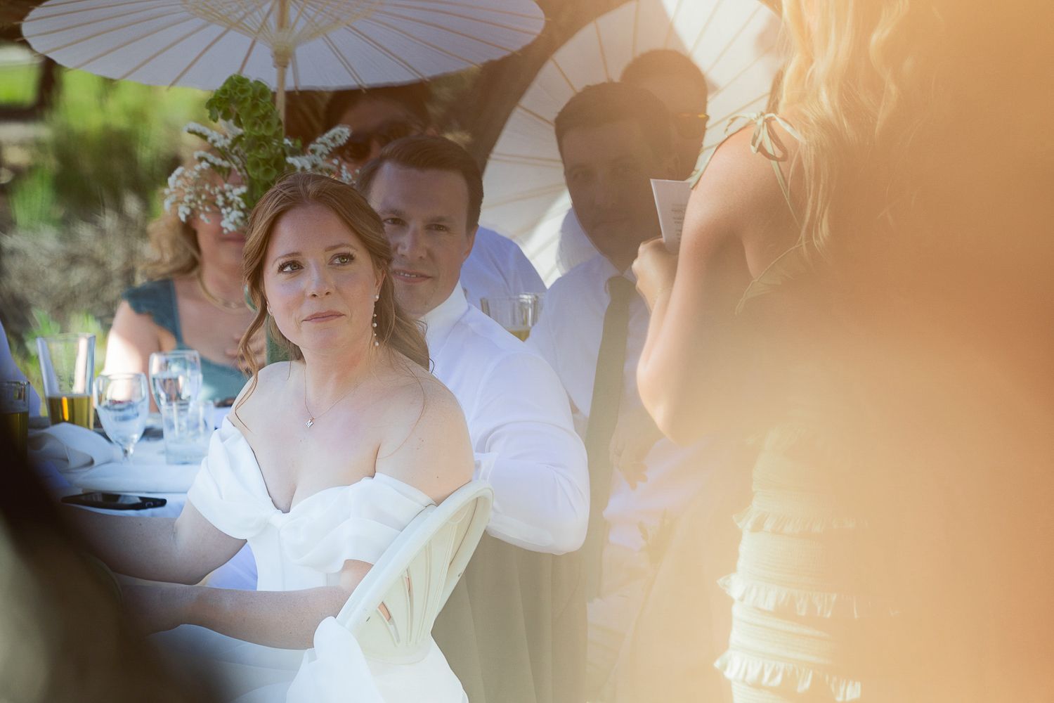 Guests seated at an outdoor wedding reception under white parasol umbrellas in soft lighting.