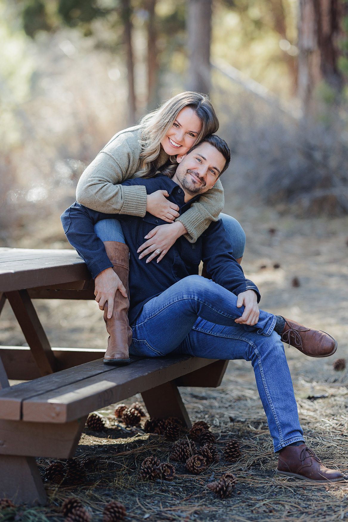 A couple shares a tender embrace while sitting on a rustic wooden bench in a forest setting with pinecones scattered below.