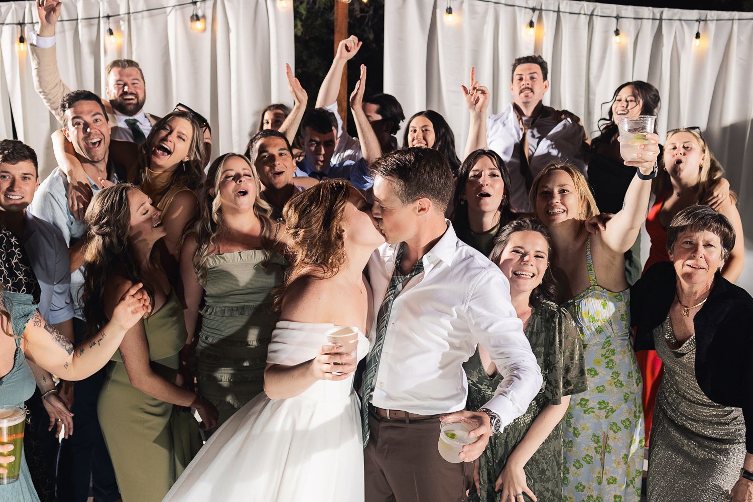 Wedding reception crowd celebrates enthusiastically as newlyweds embrace during lively dance party indoors.