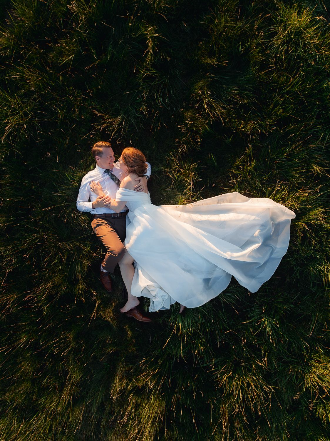 Aerial view of a couple lying in tall grass, with a flowing white wedding dress creating an elegant contrast against the green.