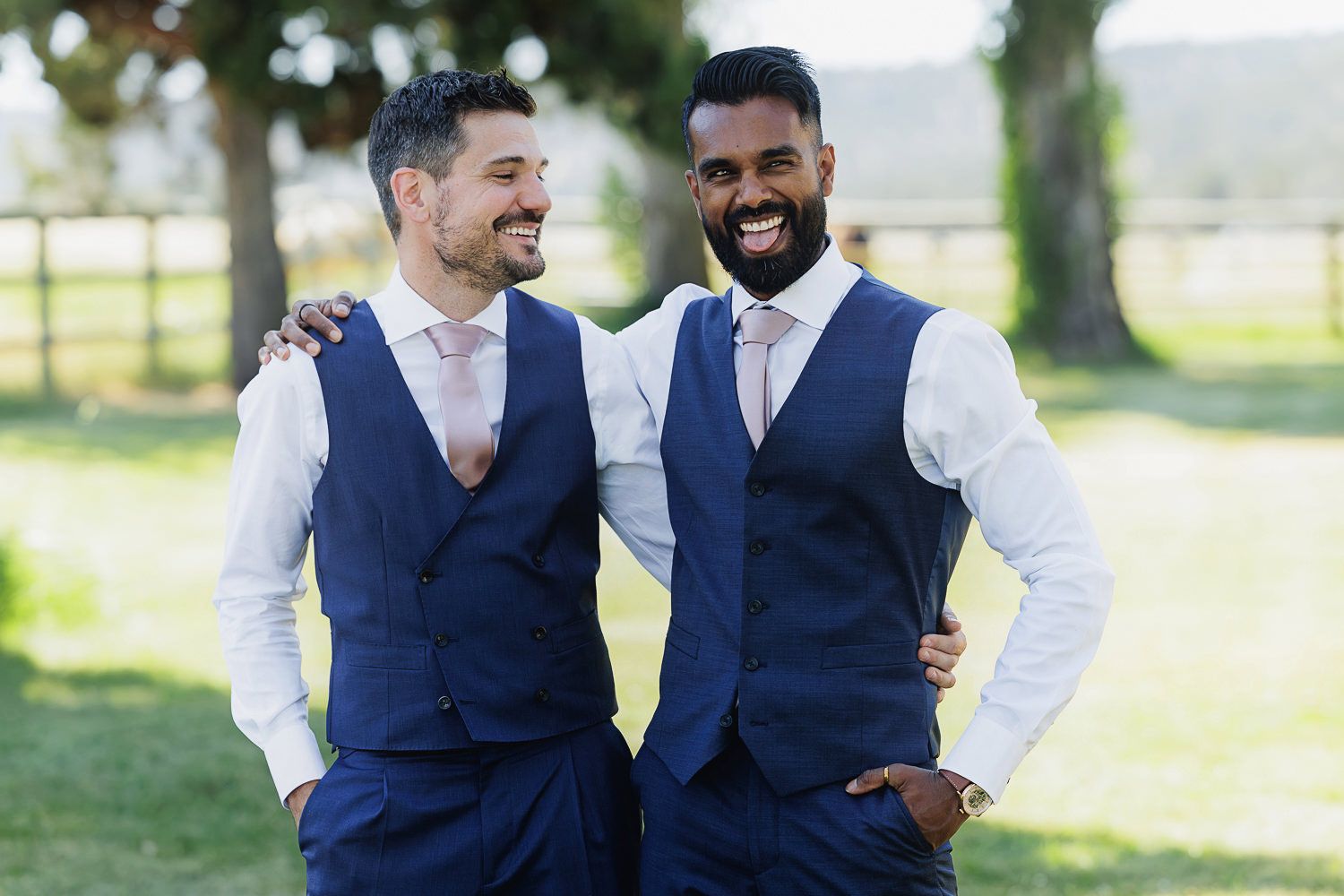 Two men in navy blue vests and white shirts laugh together in a sunny outdoor setting.