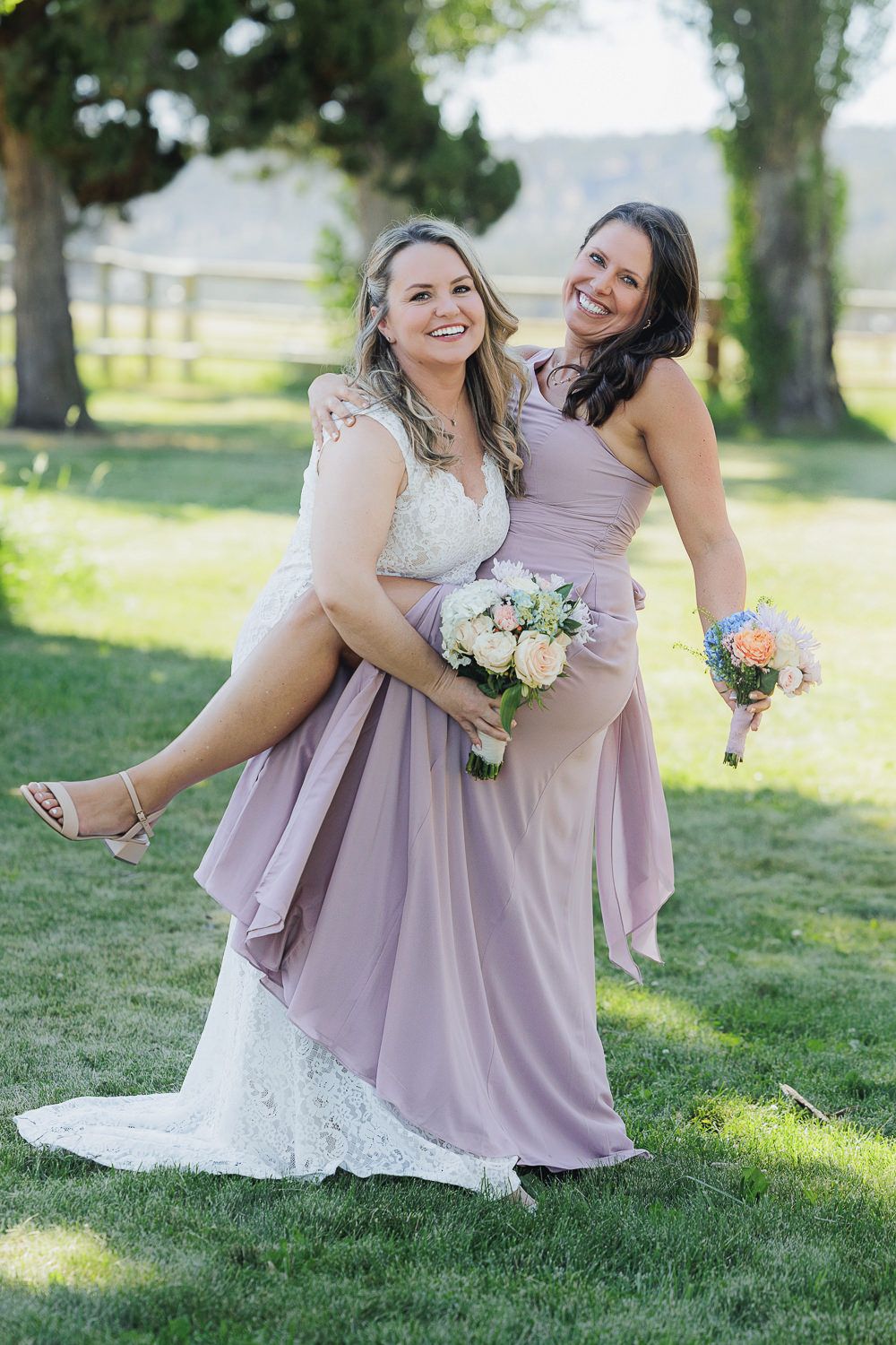 Two women in wedding attire share a playful moment outdoors, with one being carried in a fun pose.