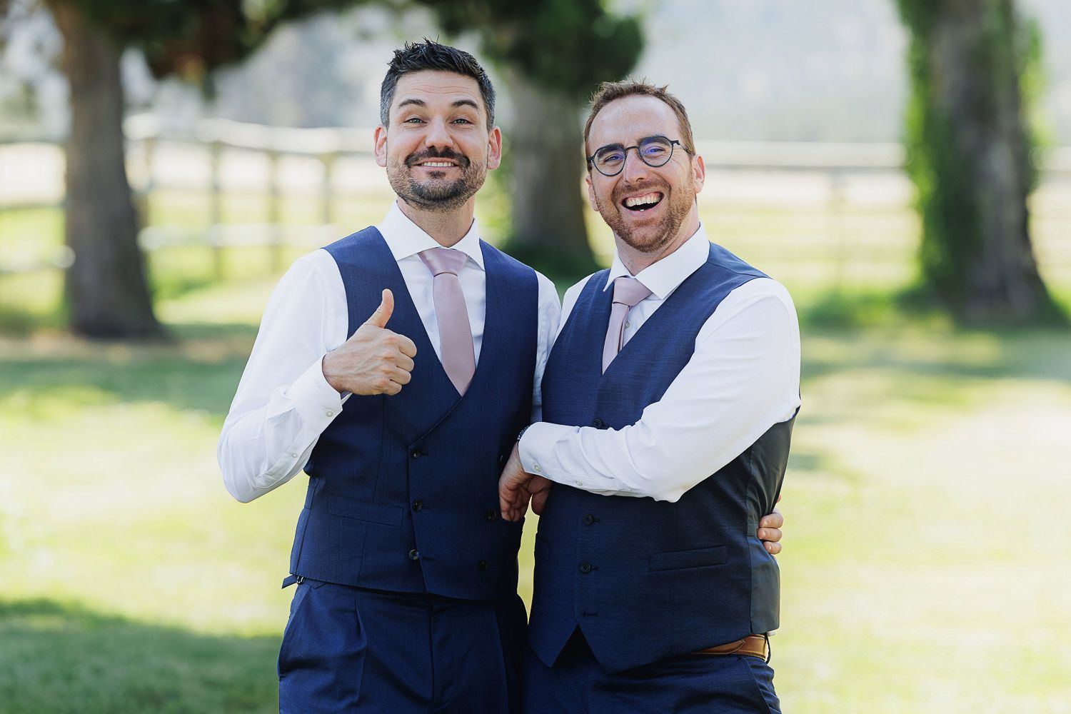Two people in matching navy blue vests and white shirts smile outdoors with white fence in background.