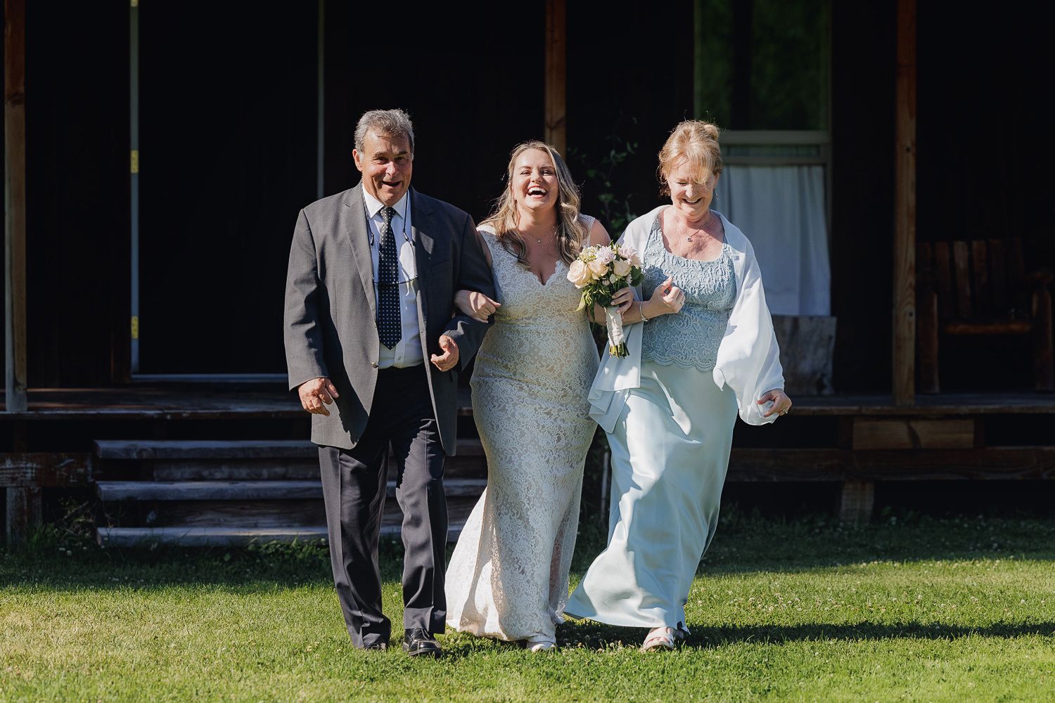 Three wedding attendees walk together on a grassy lawn outside a rustic wooden building on a sunny day.
