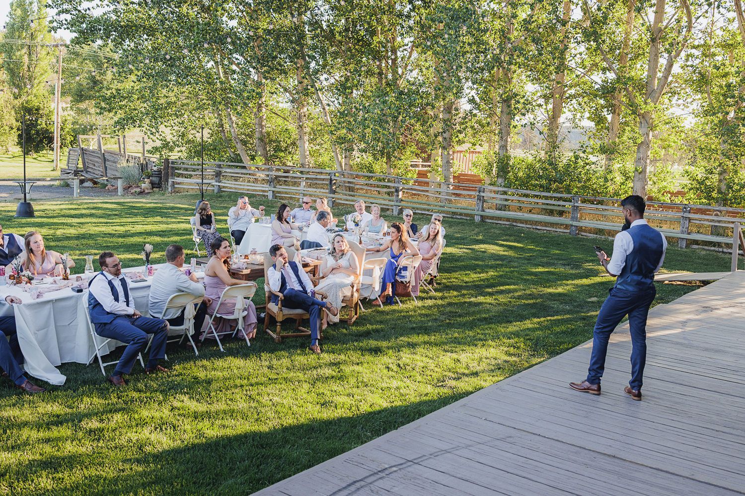 Outdoor wedding reception on a grassy lawn with guests seated at long tables and someone speaking from a deck.