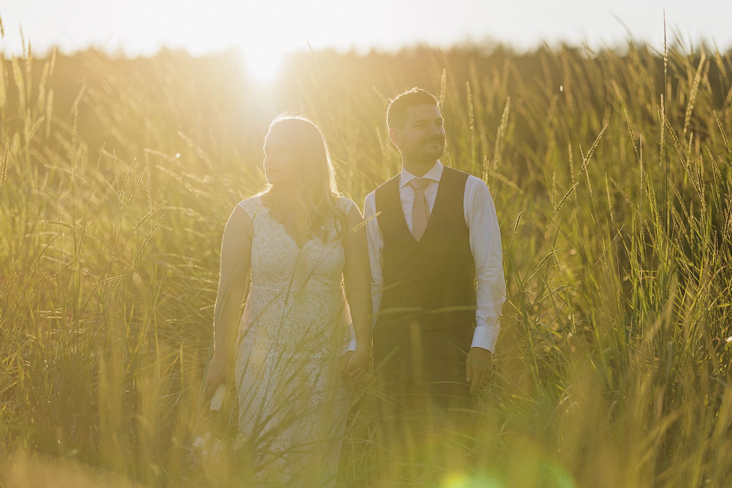A romantic couple stands together in tall grass during golden hour sunset.