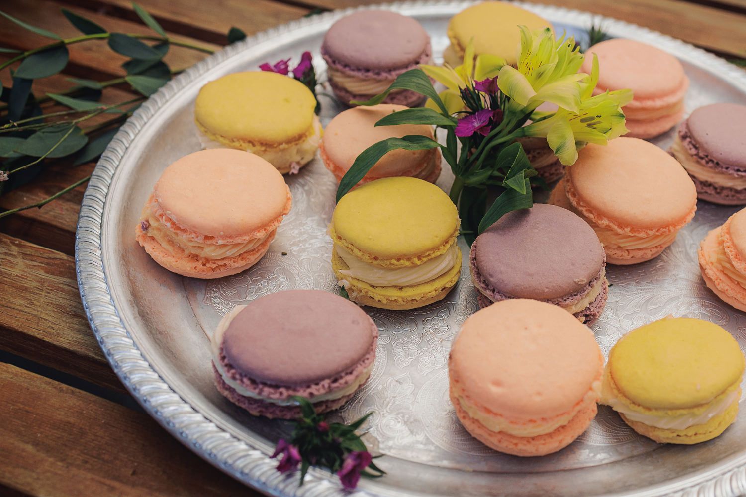 Colorful French macarons in pastel shades of yellow, pink, and purple arranged on a white decorative plate with flowers.