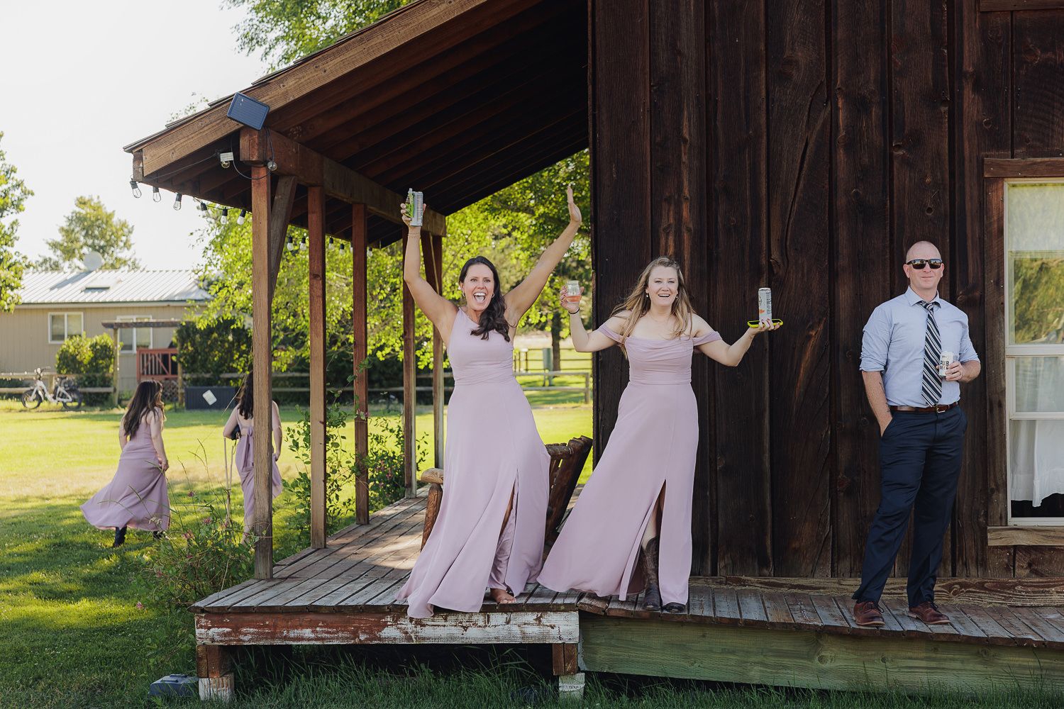 Wedding party members in light pink dresses pose playfully on a rustic wooden porch during an outdoor celebration.