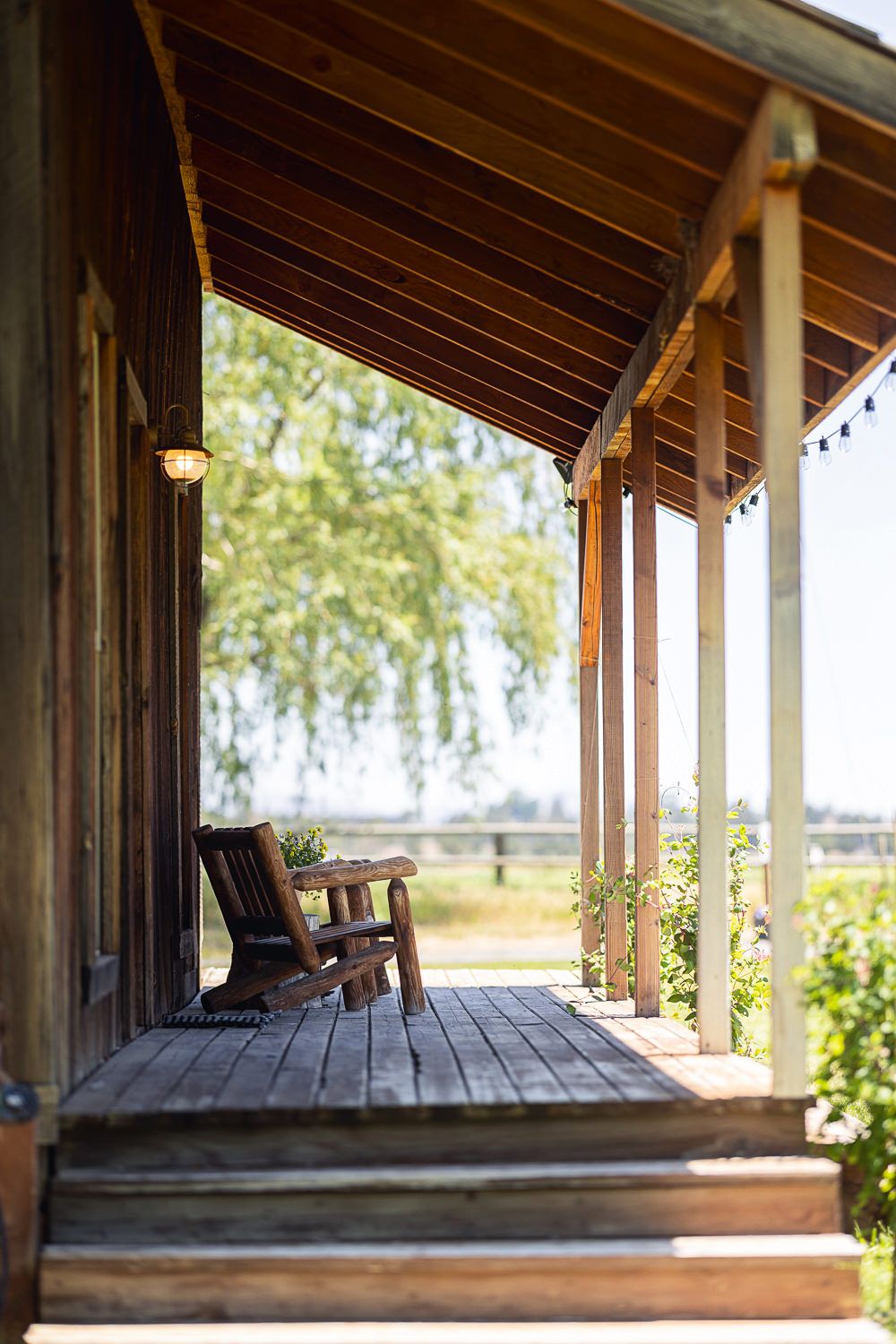 Rustic wooden porch with rocking chair overlooking sunny countryside landscape through exposed beam roof structure.