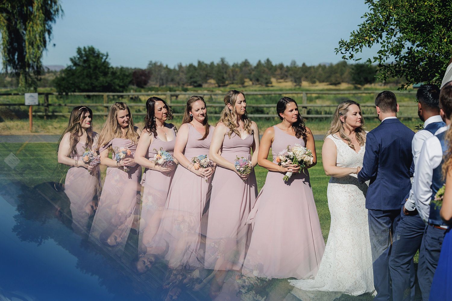 Wedding party lined up outdoors wearing pink bridesmaid dresses and navy attire for a photo by a lake.