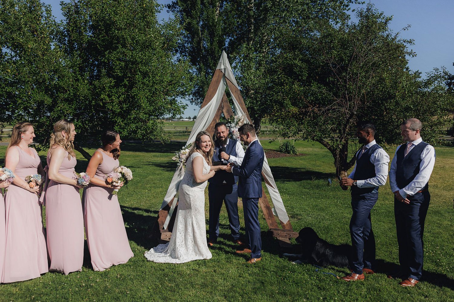 Outdoor wedding ceremony under blue skies with attendants in pink dresses and wooden arch backdrop.