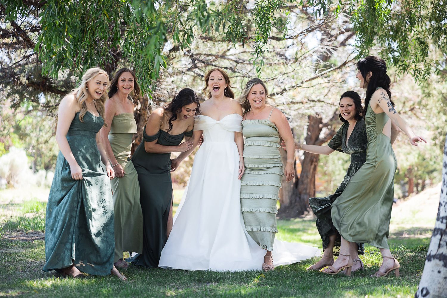 Bridal party in sage green dresses laughing and dancing together outdoors under trees on a sunny day.