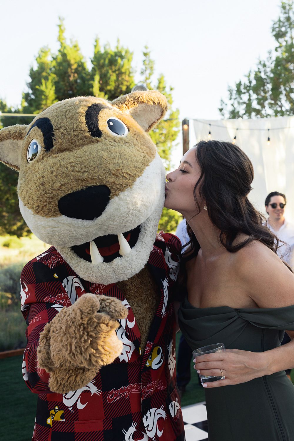 A woman in a dark dress gives a friendly kiss to a large tan and black mascot wearing a red patterned shirt.