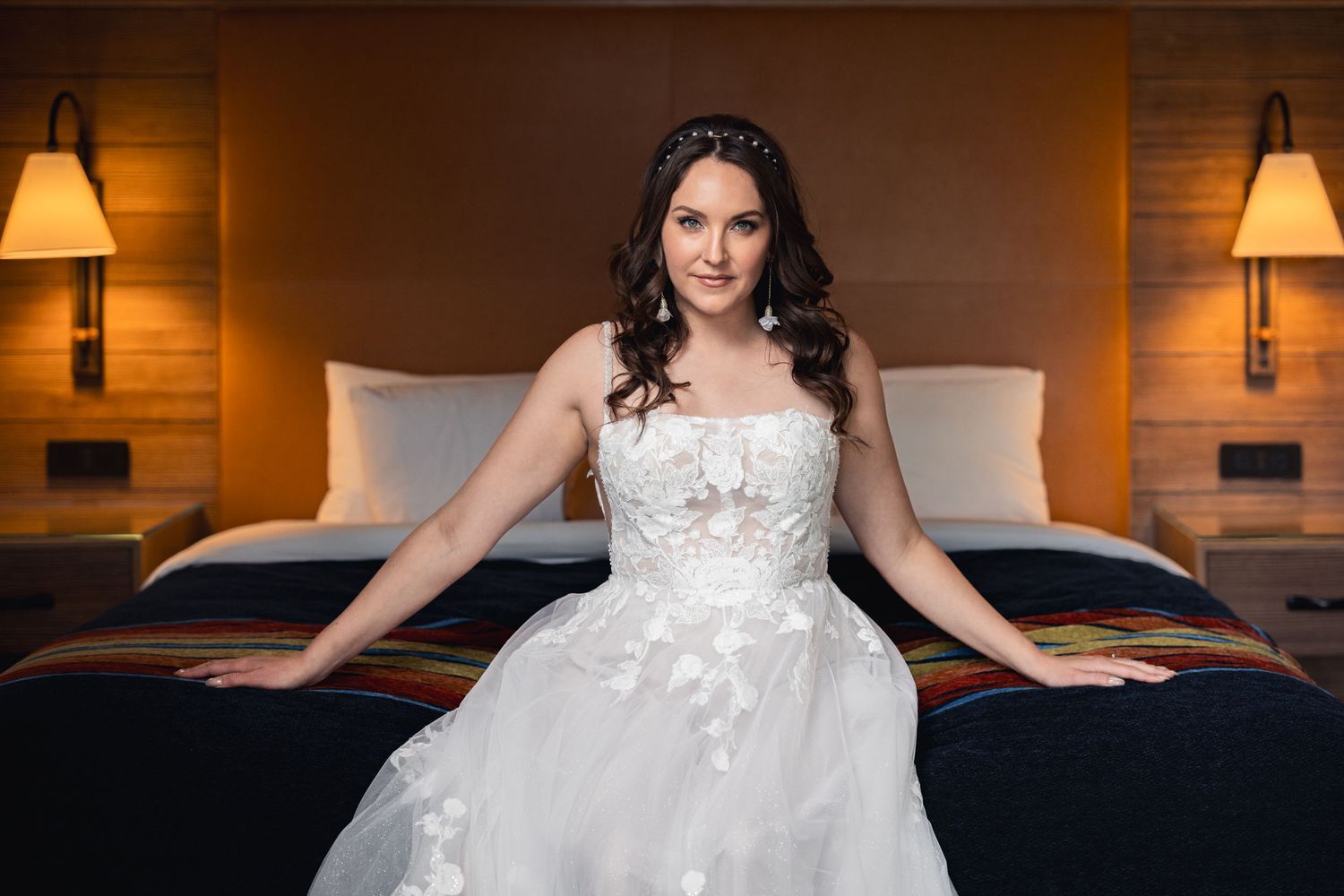 A bride in a white lace wedding dress sits elegantly on a hotel bed.