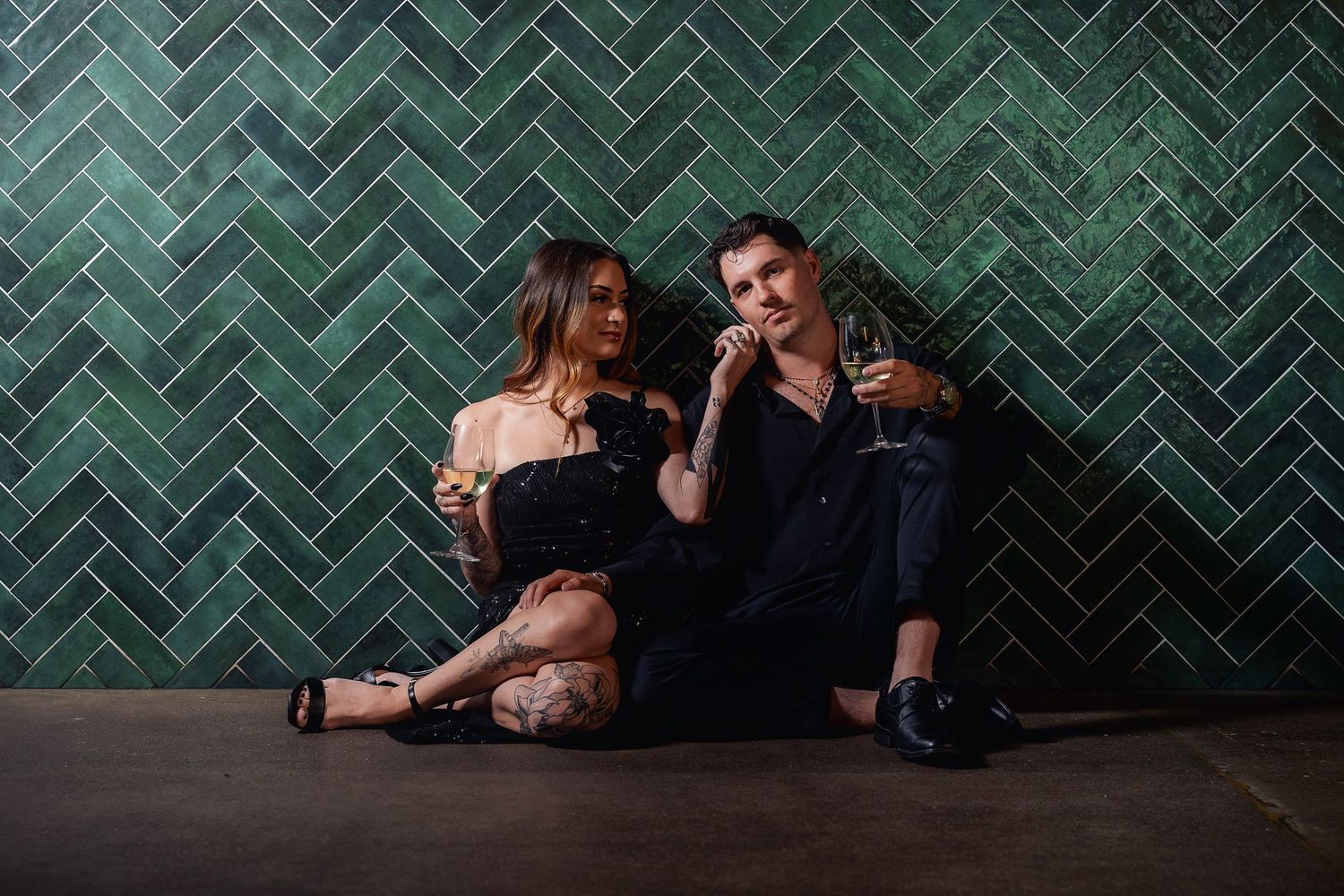Two figures sit casually on the floor against a green herringbone tile wall, holding drinks and engaging in conversation.