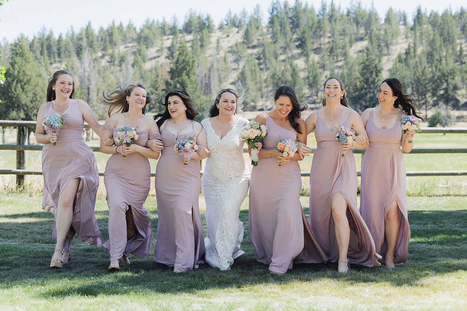 A celebratory group wearing matching mauve dresses poses outdoors on a sunny day near evergreen trees and grass.