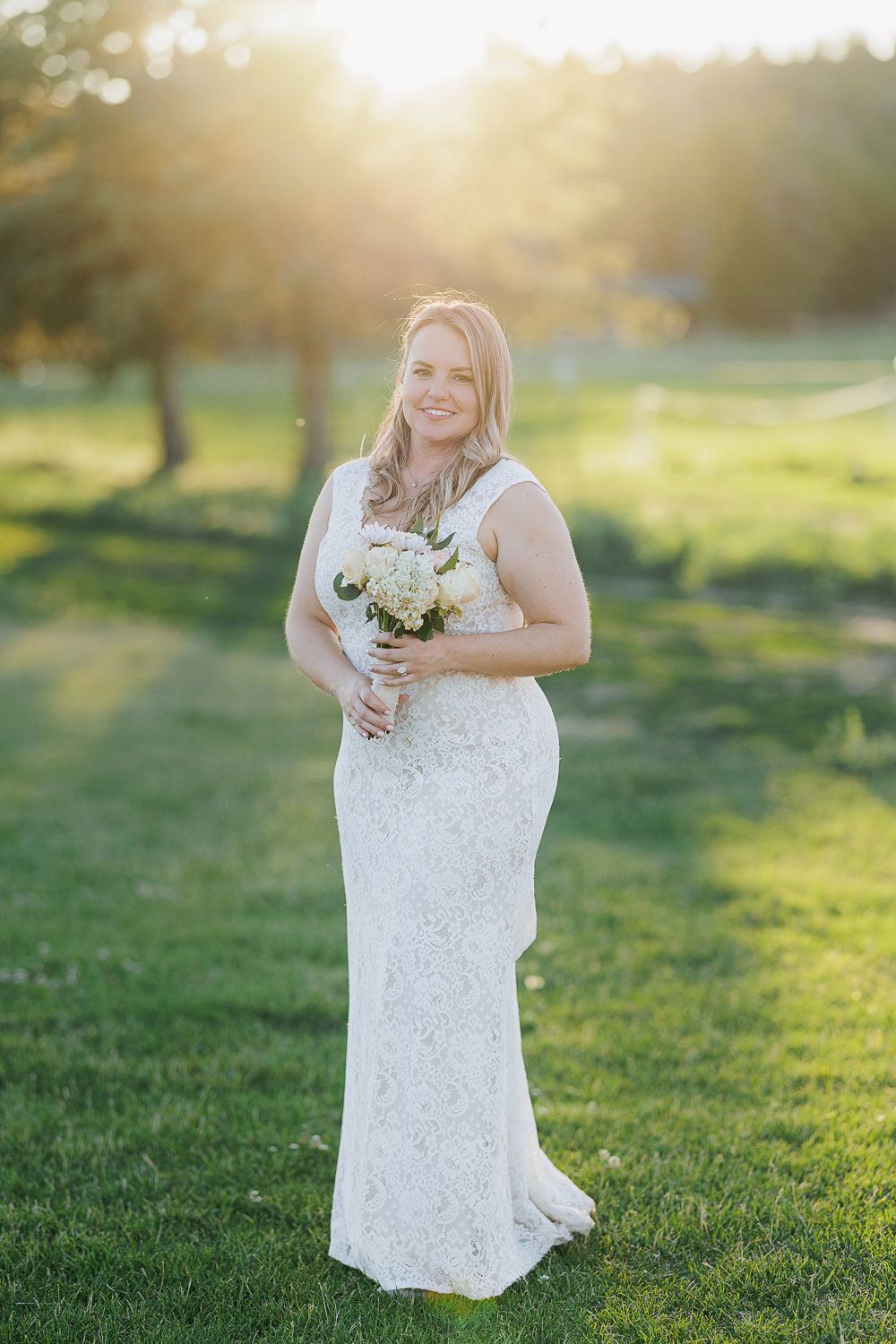 A bride wearing a fitted lace wedding gown holds a white bouquet while standing in a sunlit meadow at golden hour.