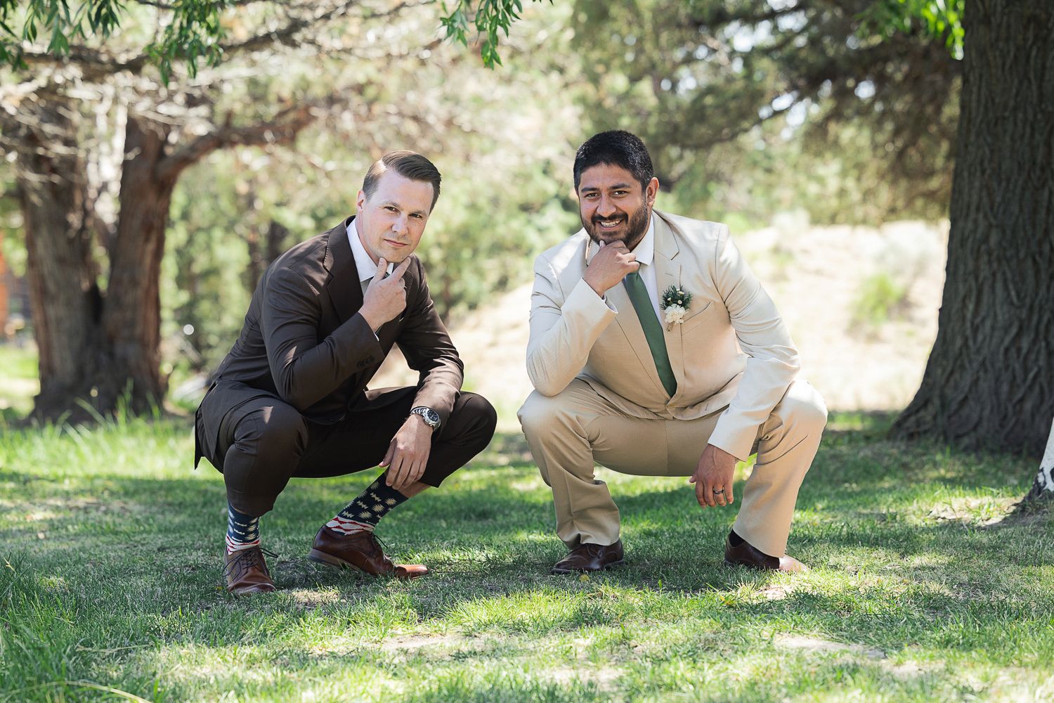 Two happy people in suits crouch together outdoors on grass against a backdrop of trees.