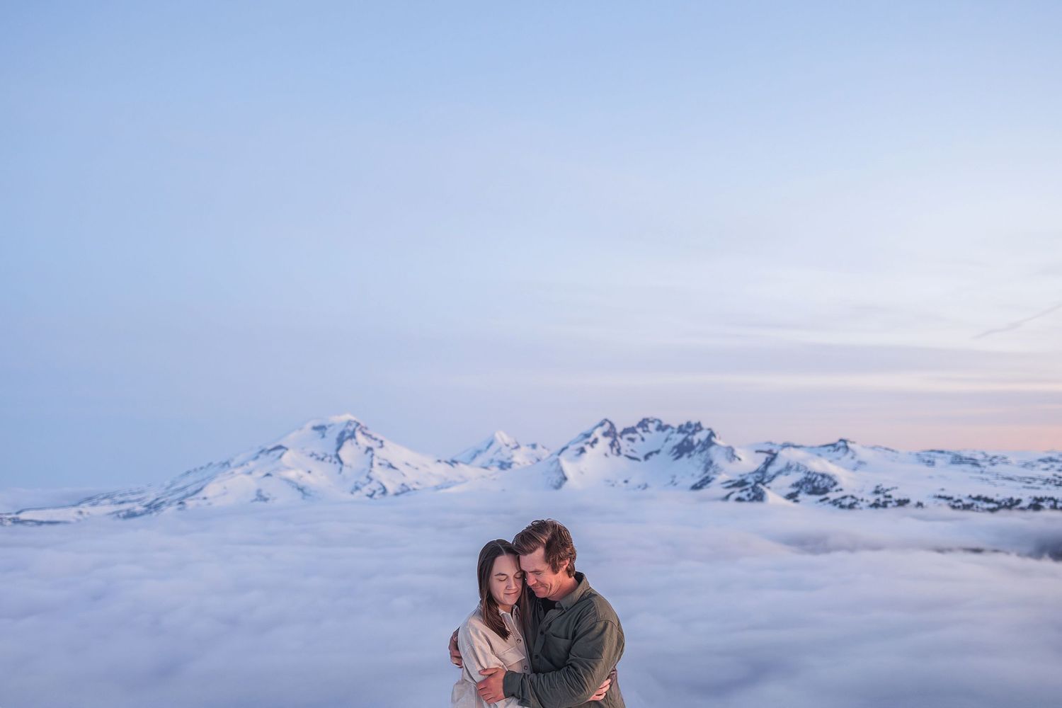 Two people embrace while standing above a dramatic cloud-covered mountain landscape at sunset.