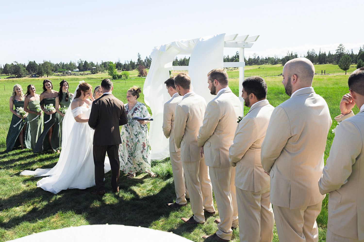 Wedding party dressed in tan and white attire gathers together for photos against a backdrop of green rolling hills.