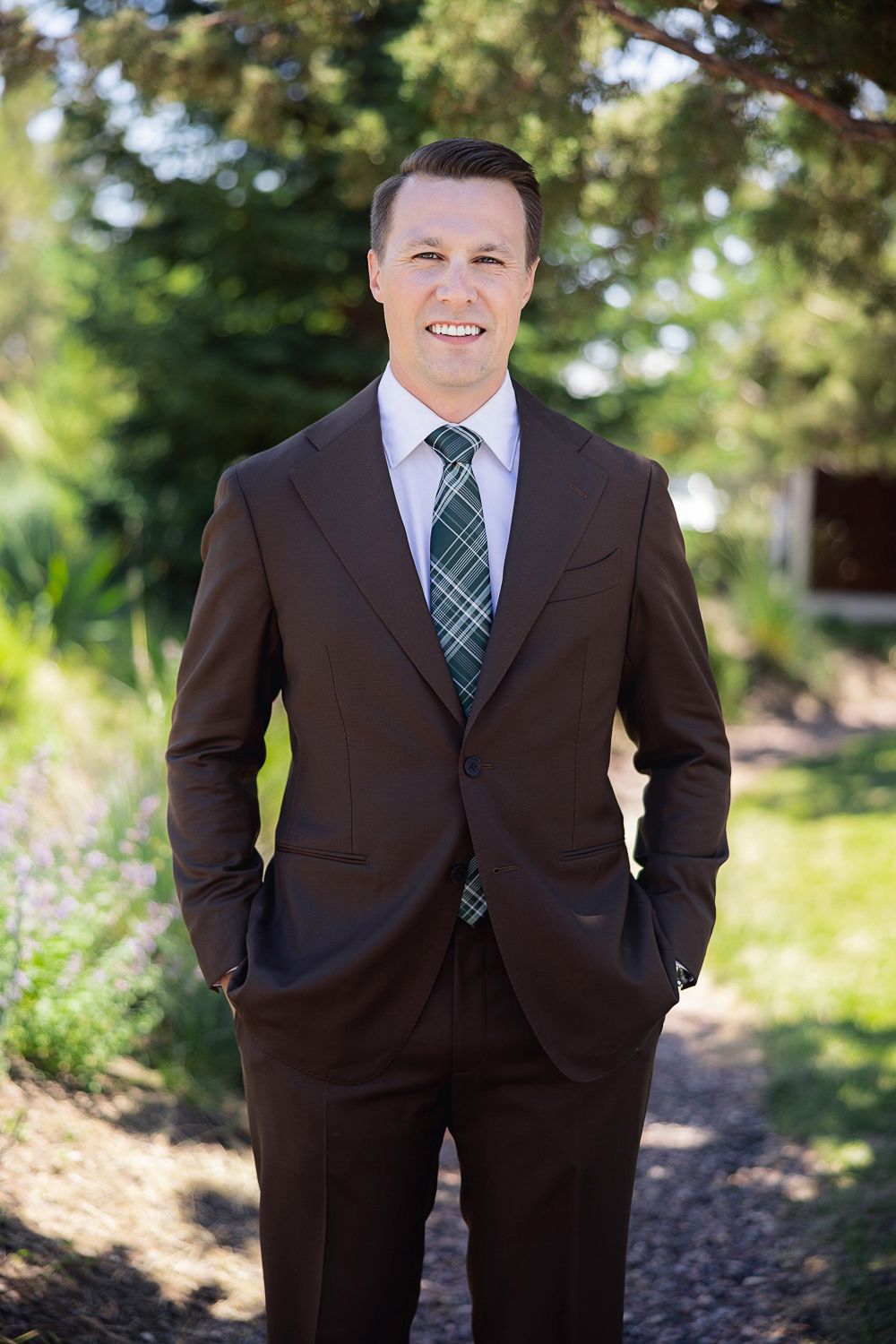 A person in a dark brown suit with a green and navy plaid tie poses outdoors in a garden setting.