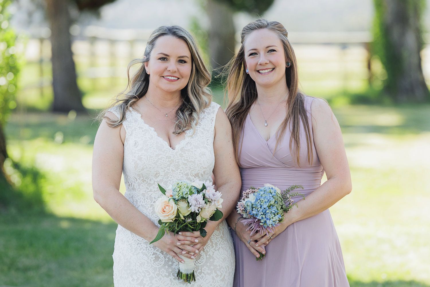 A bride in white lace gown and bridesmaid in mauve dress hold floral bouquets while smiling outdoors.