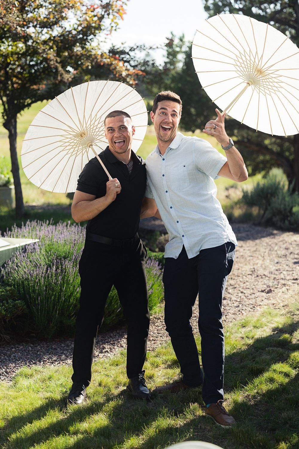 Two people posing playfully with white parasols in a sunny outdoor garden setting.