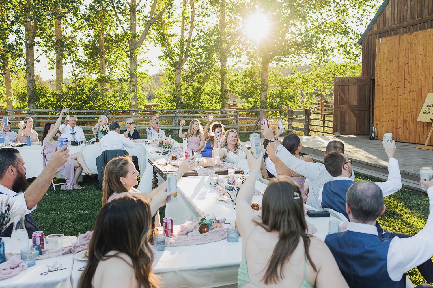 Wedding reception guests celebrate outdoors at rustic barn venue during golden hour.